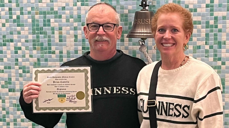 Brian Costello holds up a certificate next to a survivor's bell at Rush while standing alongside his wife.