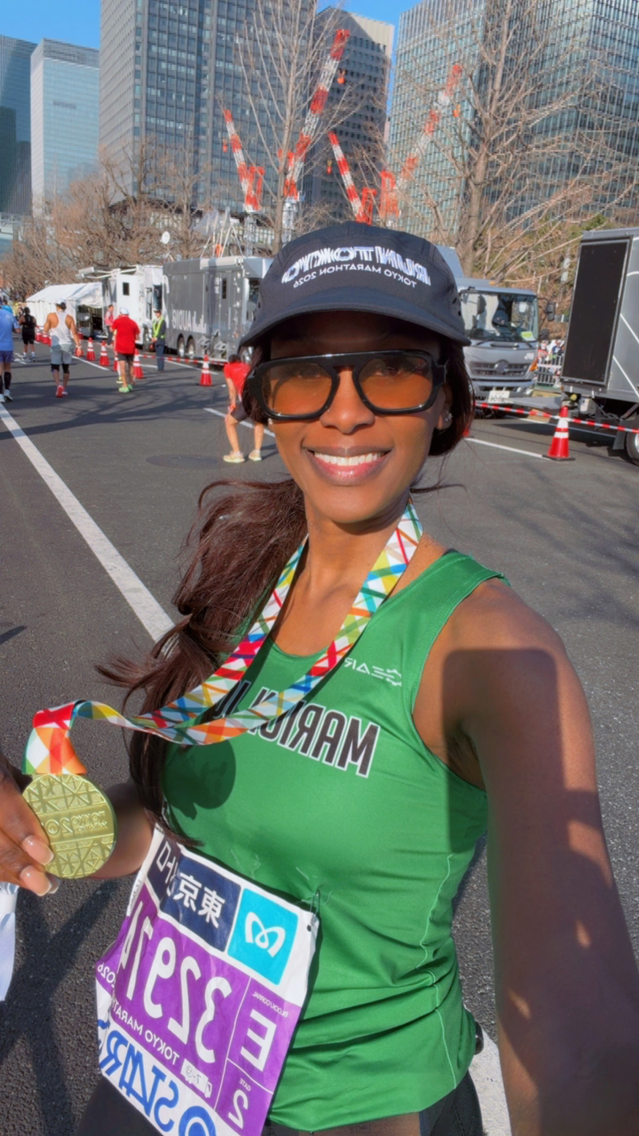 A woman in a green shirt and black hat stands on the street holding her Tokyo Marathon finisher's medal