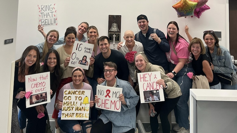 Lauren Webber stands surrounded by others, many holding supportive signs, near the survivor's bell at Rush.