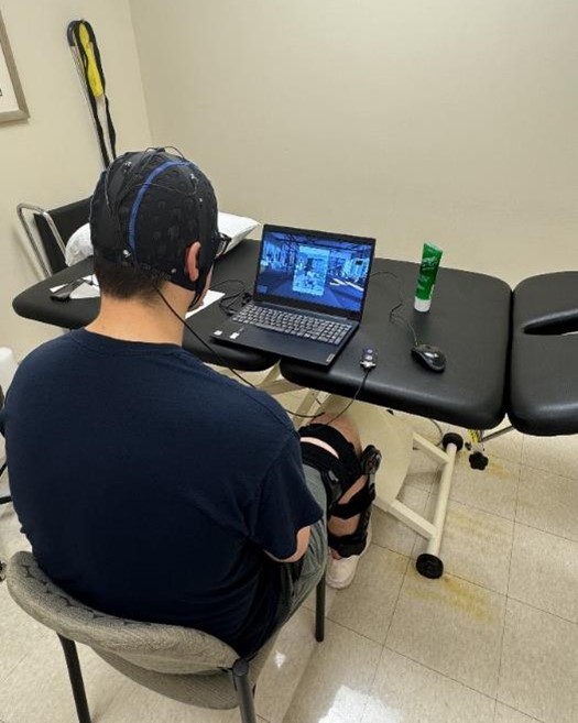 Someone sits in a chair with a brace visible on one leg and a cap on their head with wires, as they watch a screen on a laptop.