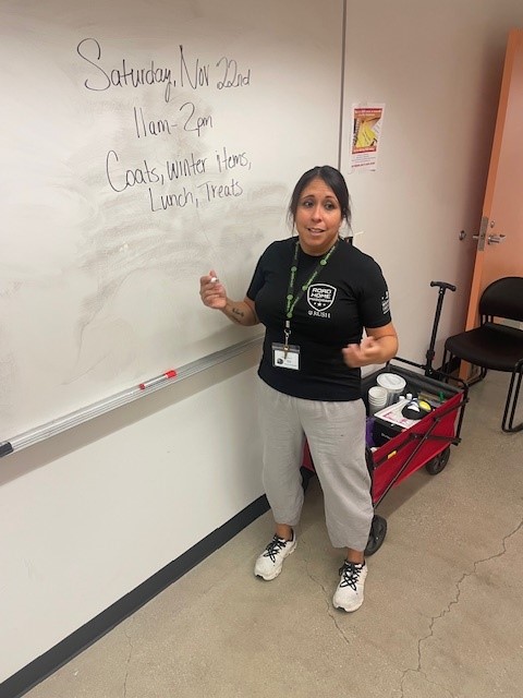 Person standing by a whiteboard with text about a Nov. 22 event from 11 a.m.–2 p.m. offering coats, winter items, lunch, and treats; a red supply cart is nearby.