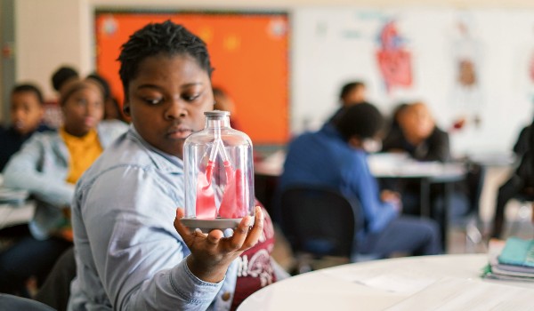 A student seated in a classroom holds a glass model representing a pair of human lungs