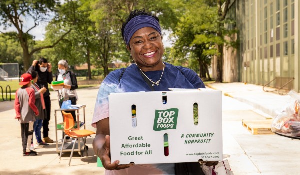 A smiling woman holds a large cardboard box that says Top Box Foods on the side