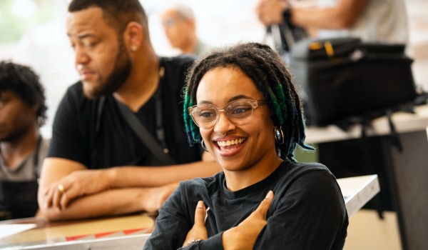 A classroom where several people are seated, with one in the center of frame wearing glasses and smiling