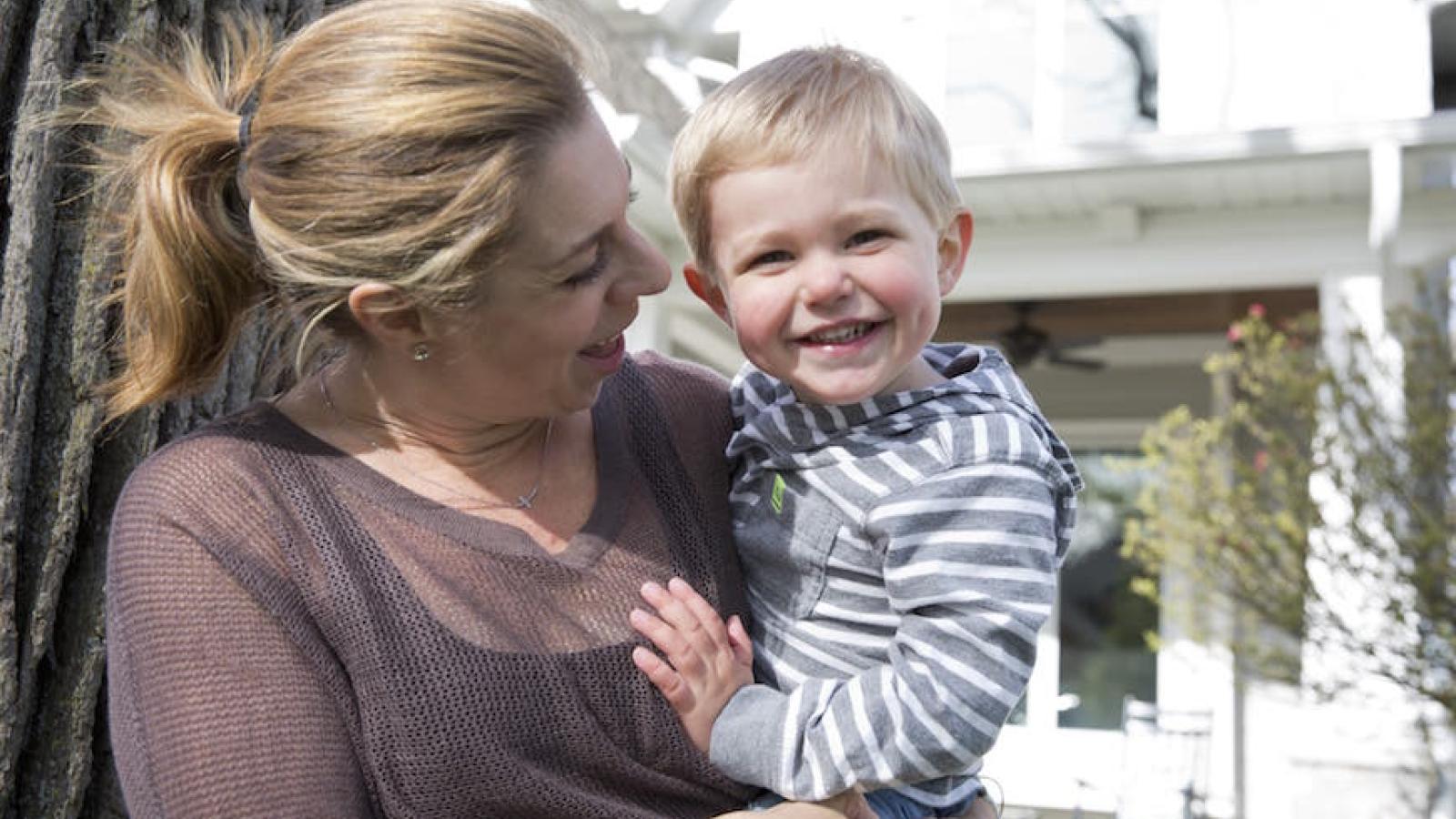 Mother smiles at her smiling toddler son while holding him outside. 