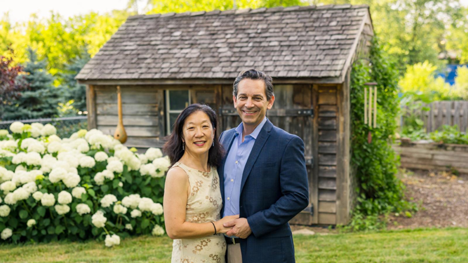 Two people standing close together in front of a rustic wooden cabin with a shingled roof, surrounded by a garden featuring white hydrangea flowers and greenery.