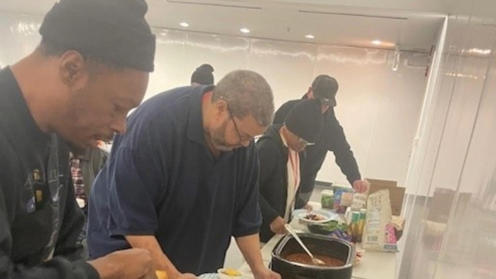 People serving themselves food from large containers on a table with plates, utensils, and packaged items.