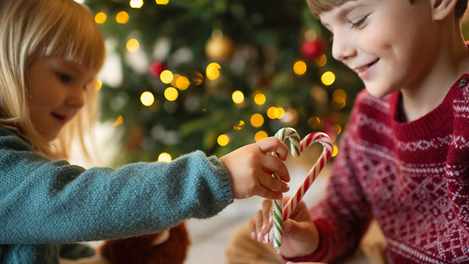 Two kids sitting under a Christmas tree hold up candy canes in the shape of a heart.