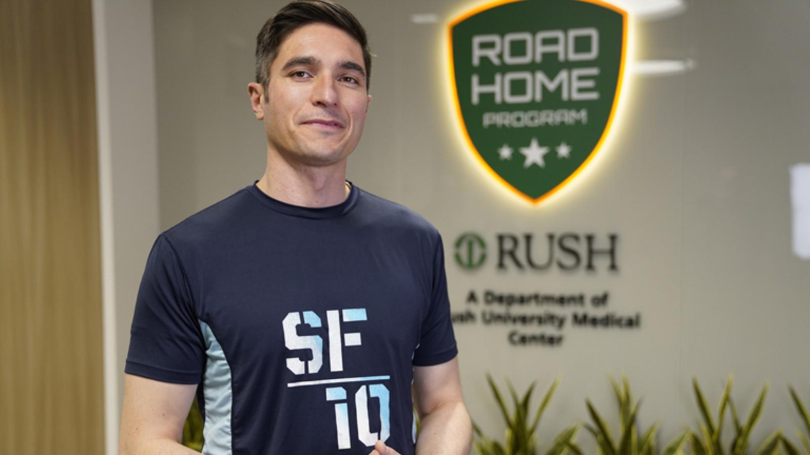 A man wearing a blue Soldier Field 10 Mile race t-shirt stands in front of a green sign that reads Road Home Program.
