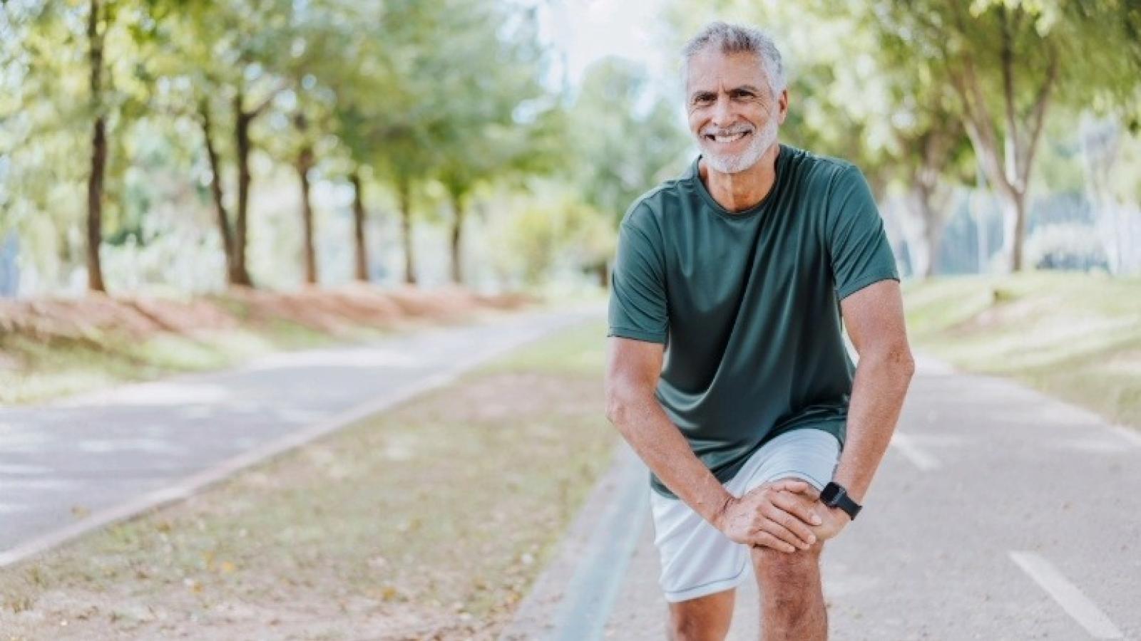 A person with short gray hair is smiling while stretching on a pathway in a park. The individual is wearing a dark T-shirt and light shorts, with trees lining both sides of the path.