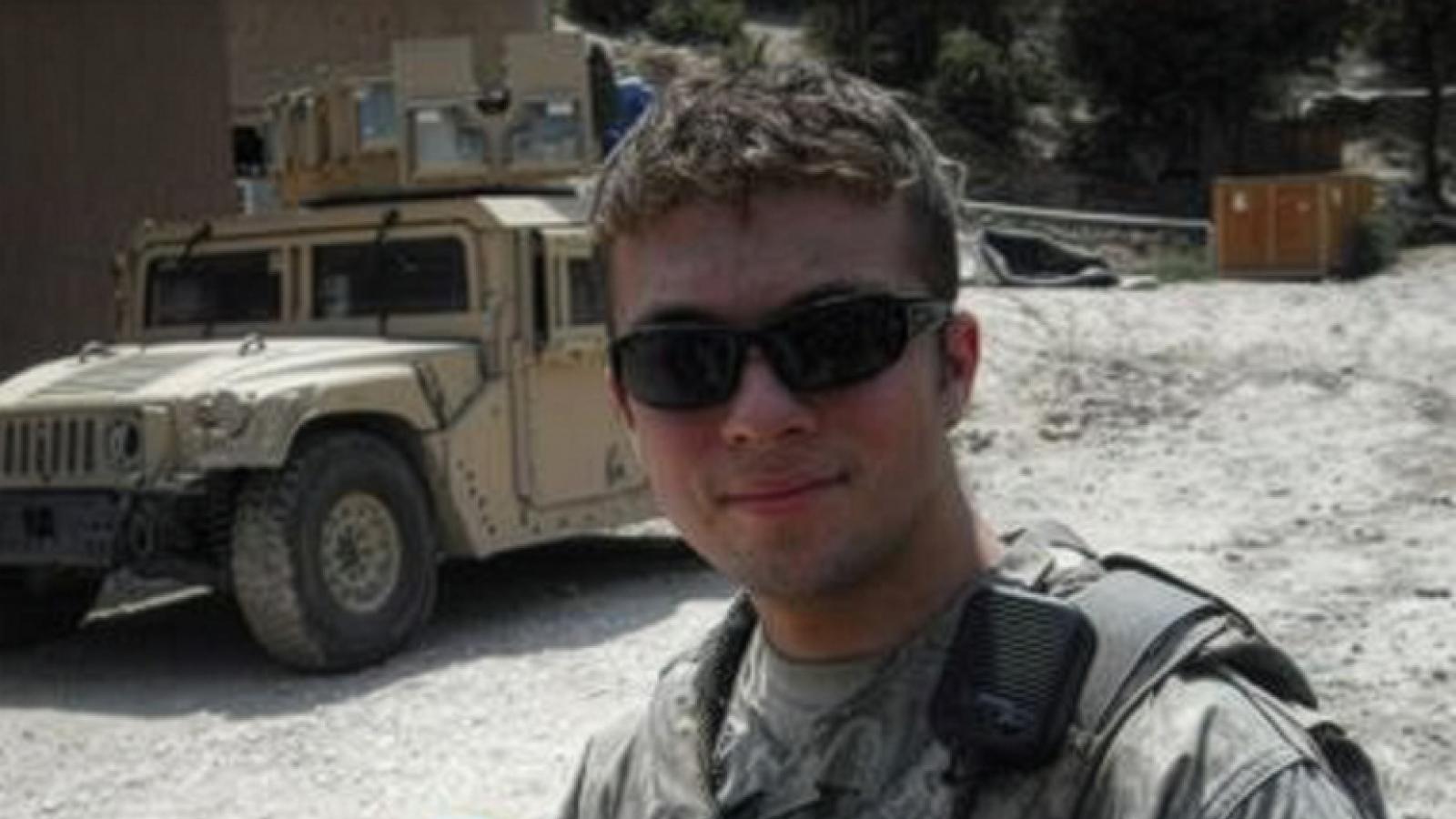 A man wearing mlitary gear and wearing sunglasses stands near a a military vehicle.