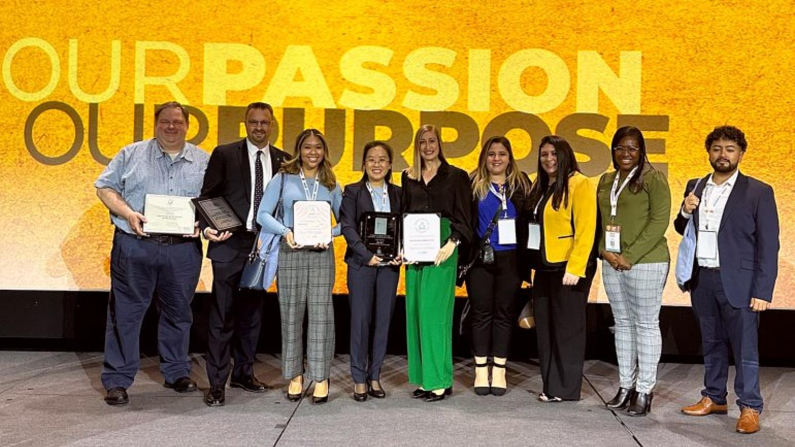 A group of students and faculty pose on a stage holding award certificates