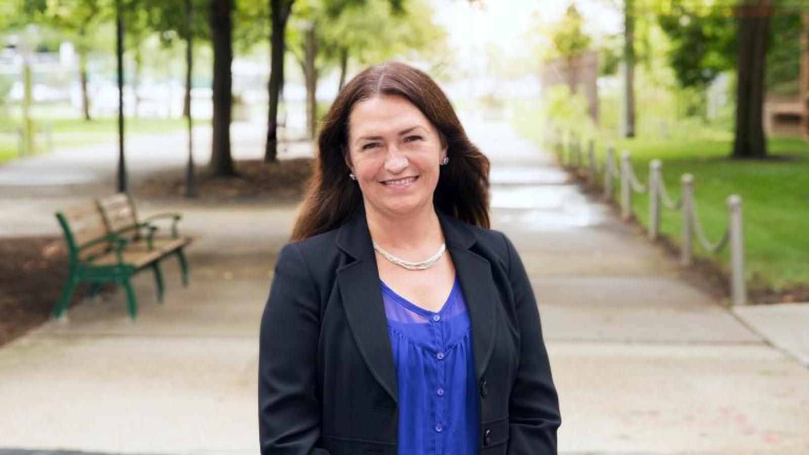 A smiling woman stands on an outdoor walkway with benches and trees 