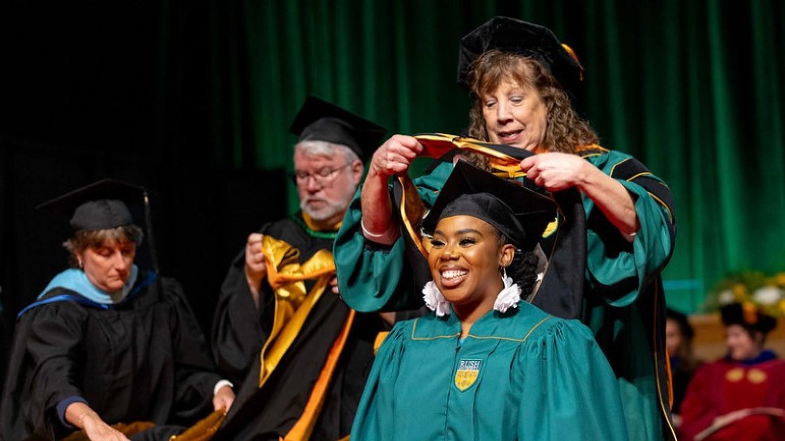 A faculty member wearing academic regalia places a sash over a smiling graduate's head
