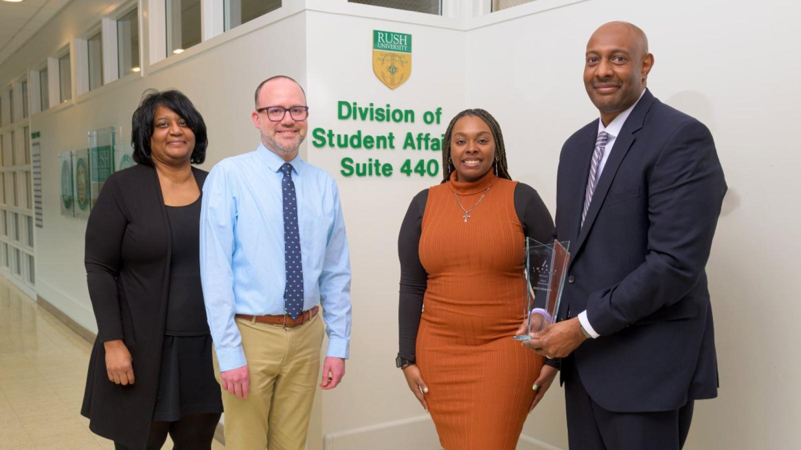 Four people standing in front of a wall sign that reads Division of Student Affairs
