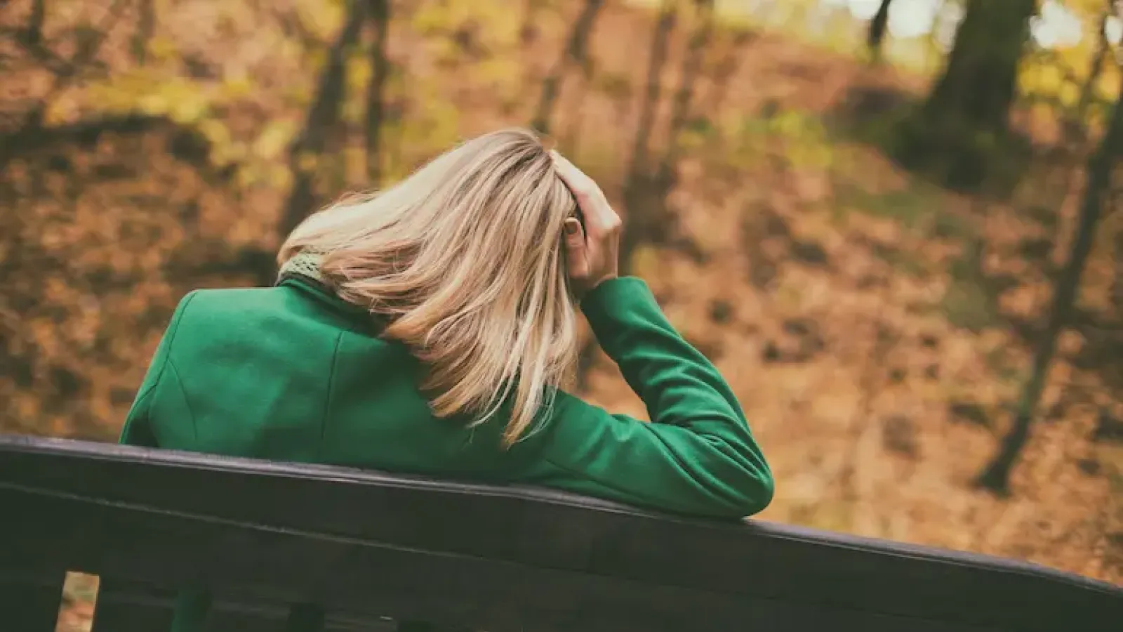 A woman in a green jacket sits on a park bench in autumn holding her head.
