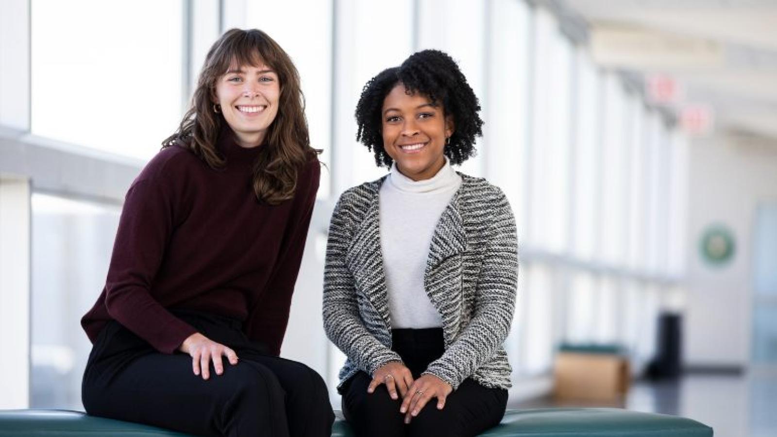 Two smiling women seated indoors on a bench in front of windows
