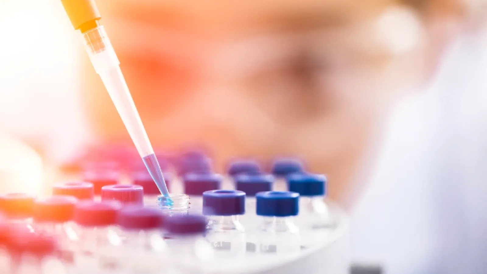 Closeup of a pipette dispensing liquid into vials, with the researcher's face wearing safety glasses out of focus behind
