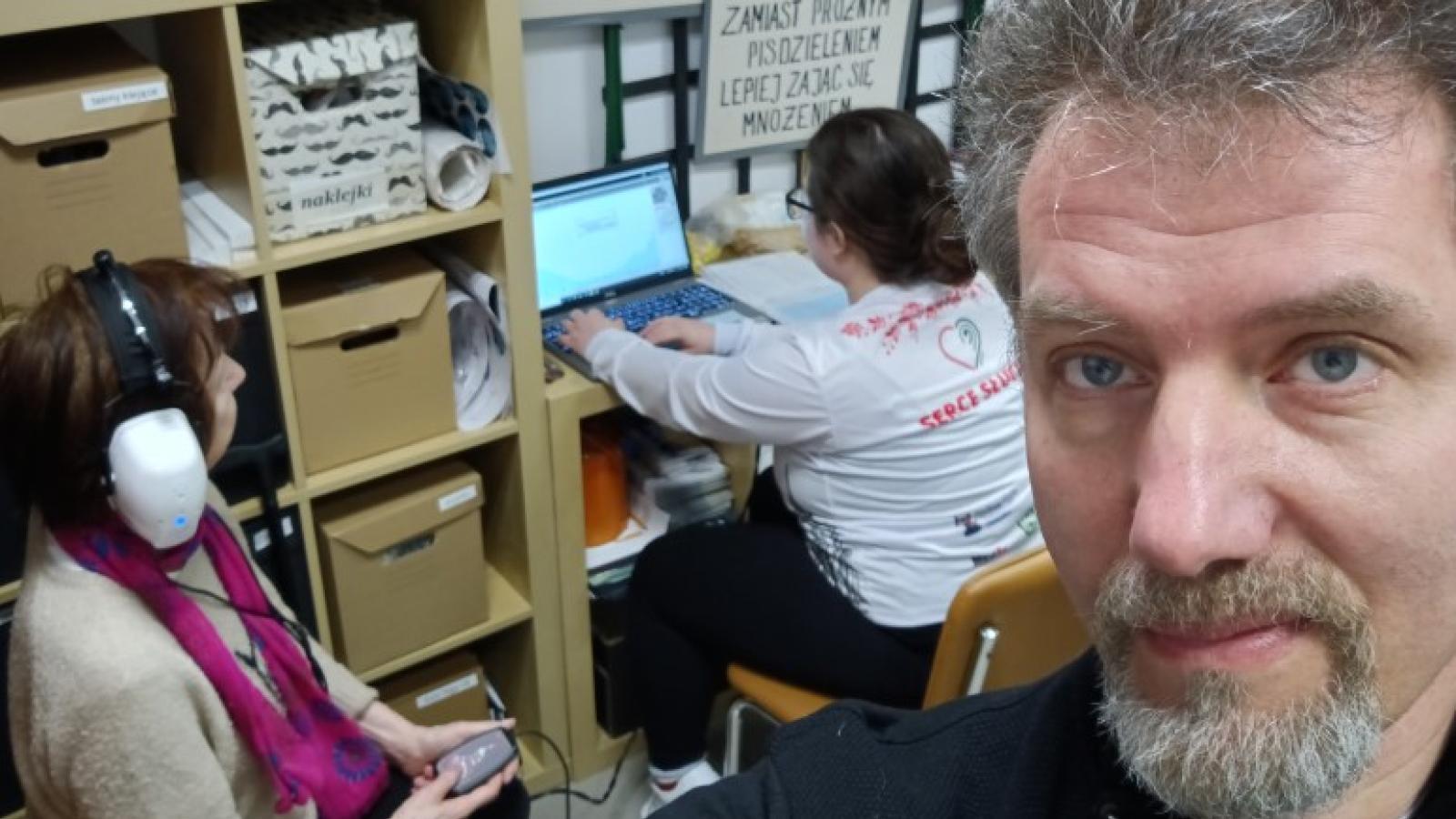 Dr. Shafiro is in the foreground as a volunteer sits at a table in the background working on a laptop that is connected to audio equipment. A patient sits to the left with headphones on, holding an audio device in her hand.