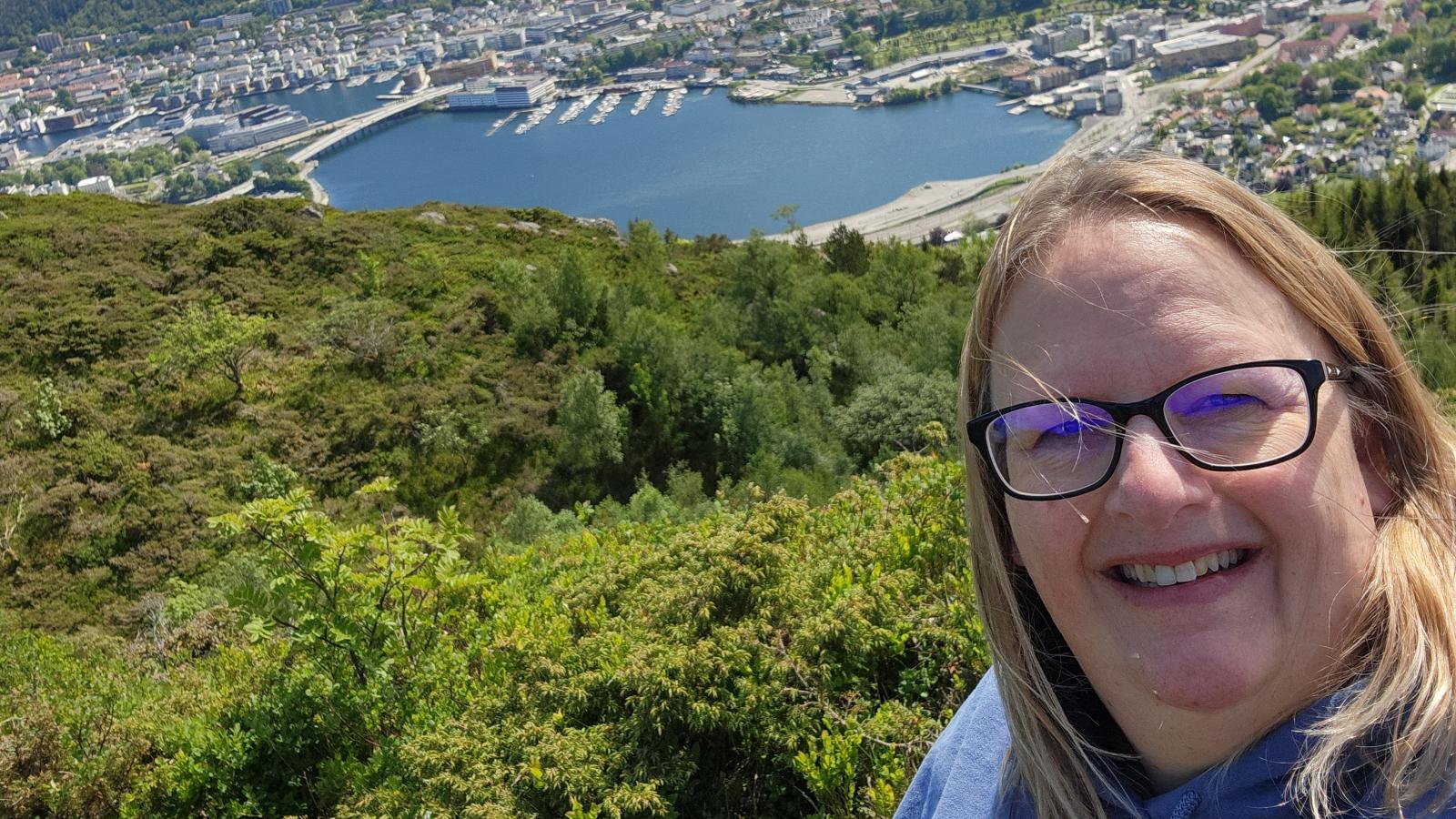 Tammy Spantiko smiles on a hill in front of a forest with a harbor and seaside city in the background.