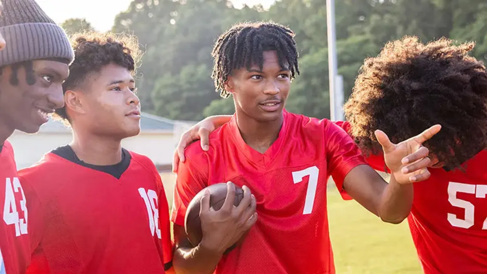 Four teen boys stand together smiling on a school football field. One is holding a football and pointing across the field.