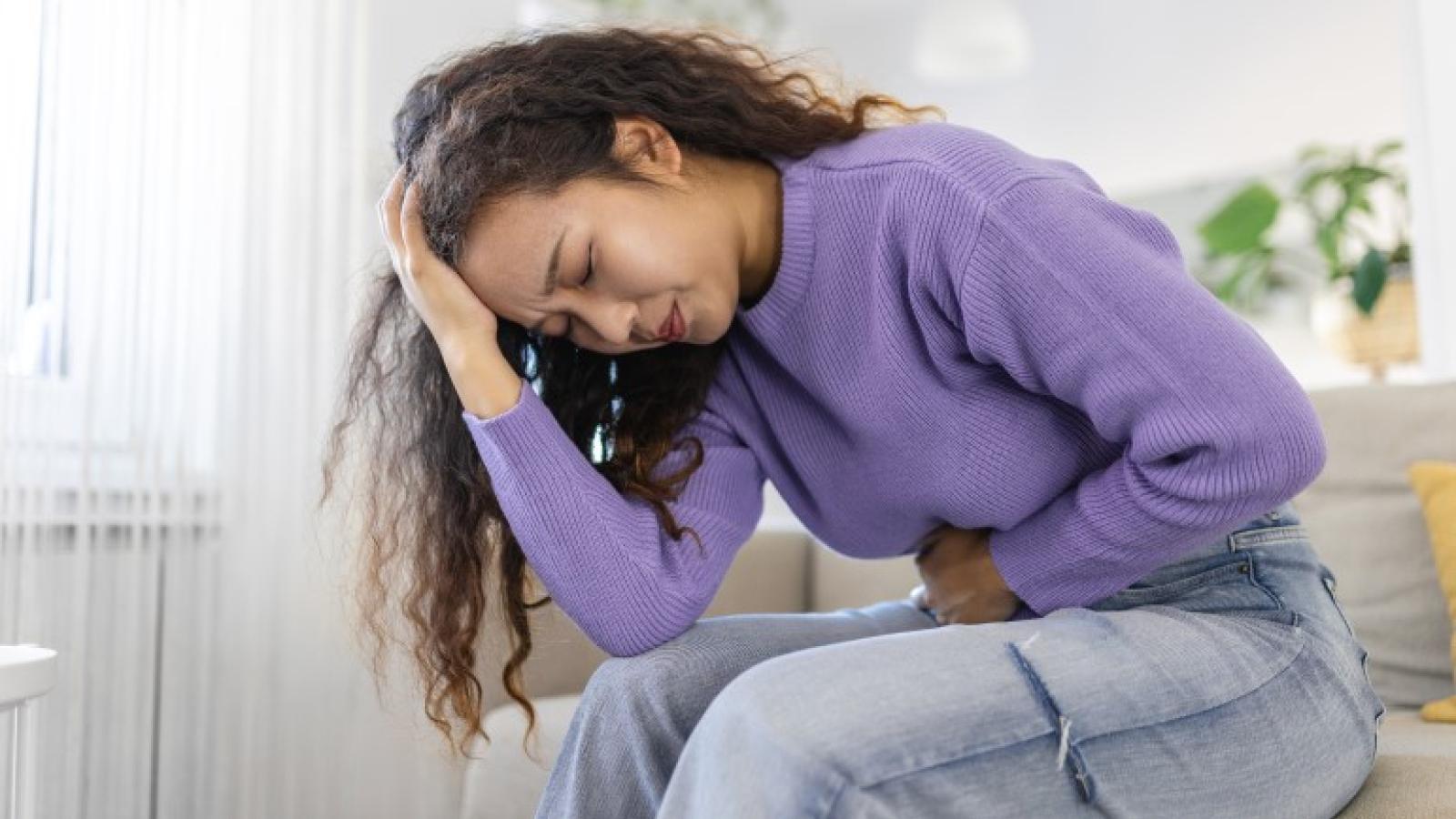 A woman sits on a sofa doubled-over in pain with one hand on her pelvic region and another on her forehead.