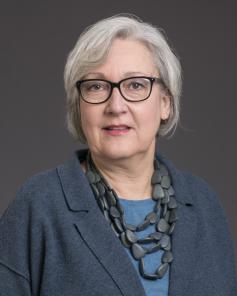 Older white woman with chin-length white hair, dark eyeglasses wearing a slate blue blazer, lighter blue shirt and a three-tier blue stone necklace. 