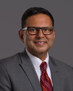 Marcos DeLeon wears a gray suit, white collared shirt, red tie and dark glasses. 