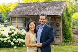 Two people standing close together in front of a rustic wooden cabin with a shingled roof, surrounded by a garden featuring white hydrangea flowers and greenery.