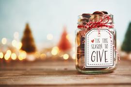 A jar decorated for the holidays reads "tis the season to give," with blurred trees and lights in the background.