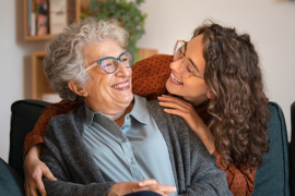 Older adult women with glasses and curly gray hair being embraced by a younger woman adult with curly brown hair.