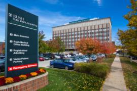Front view of RUSH Oak Park Hospital on a sunny day with blue skies. A sign in the foreground directs patients and visitors where to go for various services.