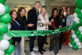 Group of people cutting a green ribbon at a grand opening event, surrounded by green and white balloon columns.
