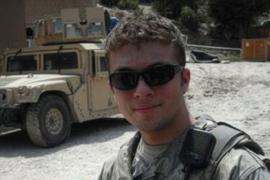 A man wearing mlitary gear and wearing sunglasses stands near a a military vehicle.