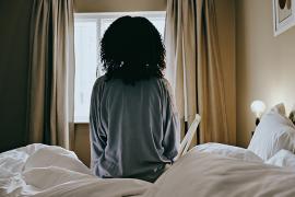 A woman sitting up in bed facing a window.