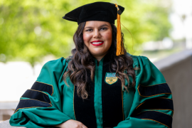 Delilah Flexas stands outdoors in full green and black commencement regalia.