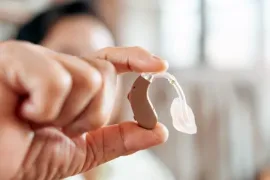 An older adult holds a behind-the-ear hearing aid to the viewer between thumb and forefinger.