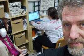 Dr. Shafiro is in the foreground as a volunteer sits at a table in the background working on a laptop that is connected to audio equipment. A patient sits to the left with headphones on, holding an audio device in her hand.