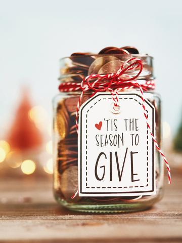 A jar decorated for the holidays reads "tis the season to give," with blurred trees and lights in the background.