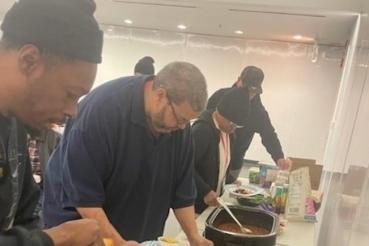 People serving themselves food from large containers on a table with plates, utensils, and packaged items.