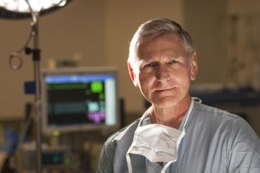 Dr. Fessler in surgical scrubs with a face mask around the neck, standing in a clinical setting with medical equipment and a monitor in the background.