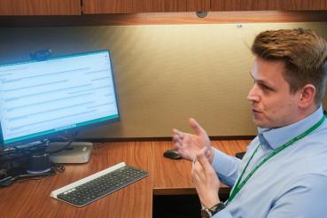A man seated at a wooden desk gesturing toward a computer monitor displaying a webpage, with a keyboard and mouse in front.