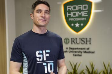 A man wearing a blue Soldier Field 10 Mile race t-shirt stands in front of a green sign that reads Road Home Program.