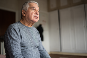 A senior Latino man looks out a window with a contemplative expression.