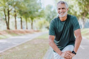 A person with short gray hair is smiling while stretching on a pathway in a park. The individual is wearing a dark T-shirt and light shorts, with trees lining both sides of the path.