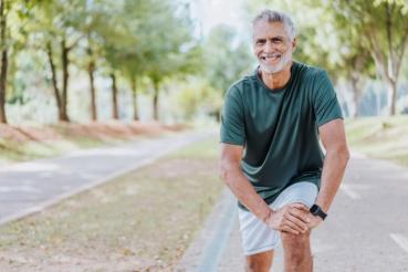 A person with short gray hair is smiling while stretching on a pathway in a park. The individual is wearing a dark T-shirt and light shorts, with trees lining both sides of the path.