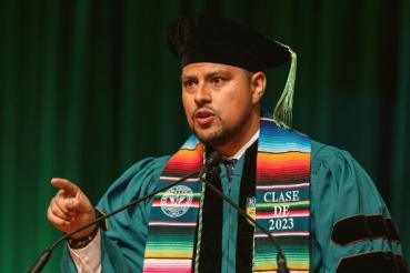 Jordan Cisneros, MD, speaks into a microphone and makes a pointing gesture with his right hand. He wears graduation regalia including a stole that says Clase de 2023