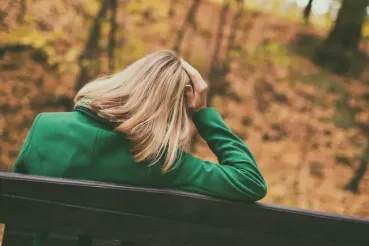 A woman in a green jacket sits on a park bench in autumn holding her head.