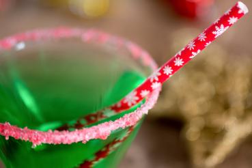 A martini glass rimmed with red and white sugar and filled with a green holiday drink. A red straw with a snowflake pattern sticks out of the glass.