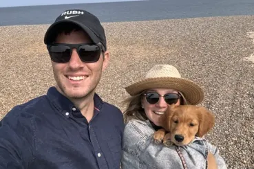 Ned Barker stands on a beach with his partner, Mandy, and their puppy.